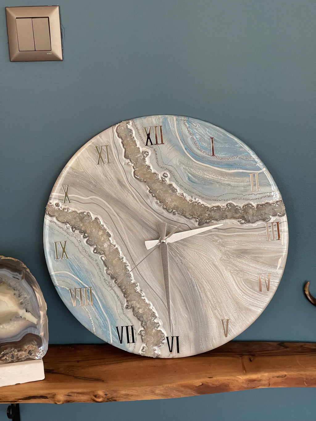 Round wall clock with a marbled design in shades of blue, gray, and white. The clock face features Roman numerals and silver-colored hands. The clock is mounted on a blue wall.