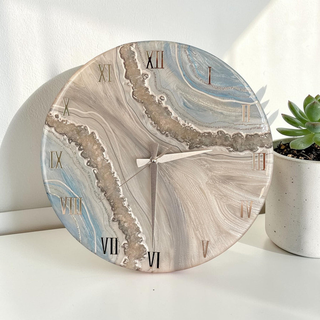 Round wall clock with a marbled design in shades of blue, gray, and beige. The clock features gold Roman numerals and hands. The clock is a decorative home decor item.