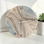 Round wall clock with a marbled design in shades of blue, gray, and beige. The clock features gold Roman numerals and hands. The clock is a decorative home decor item.