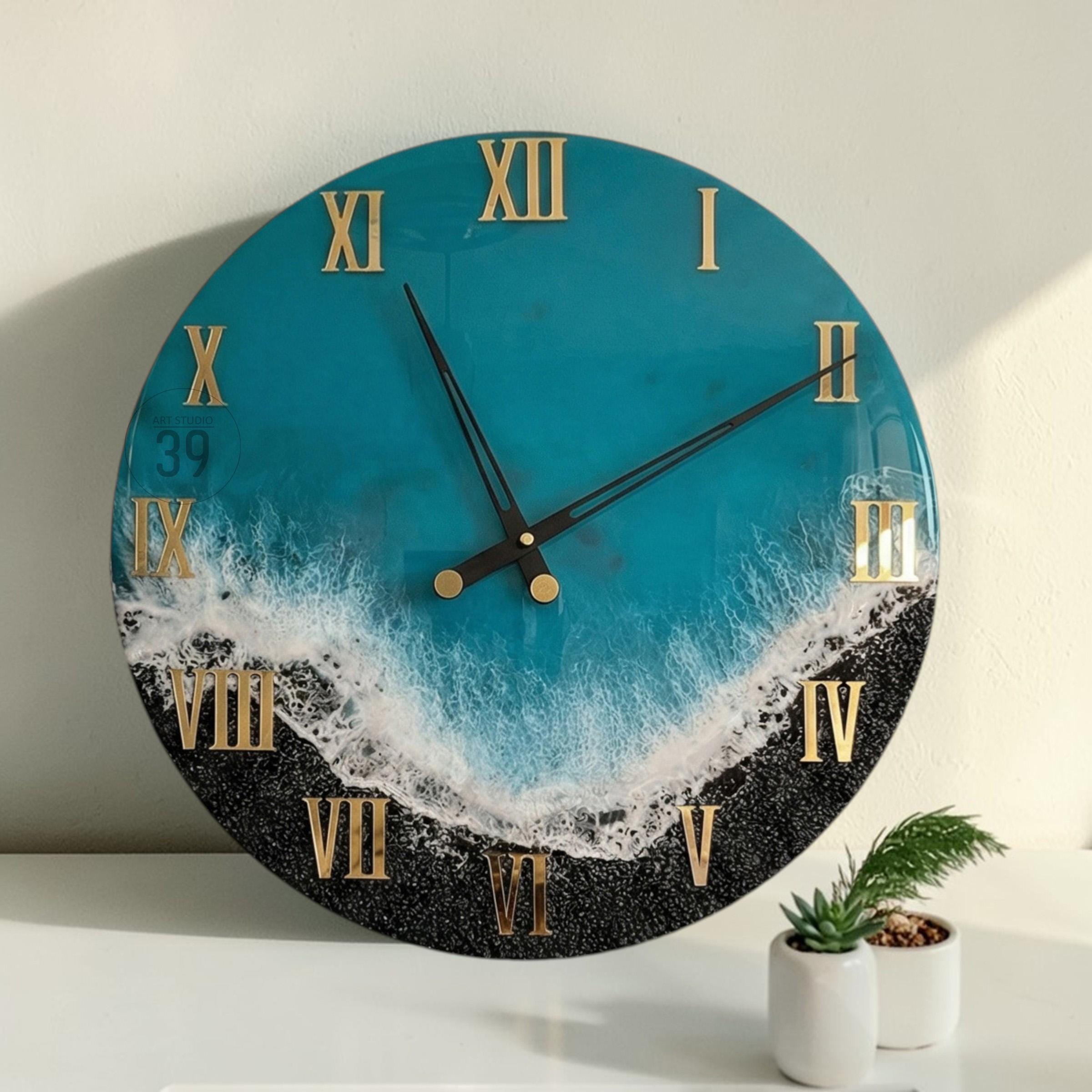 A round wall clock with a teal blue and black design, resembling a beach scene. The clock features gold Roman numerals and black clock hands. The clock is approximately 12 inches in diameter.