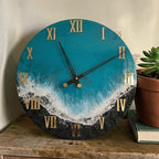 Round wall clock with a teal and black ocean-themed design. The clock face features gold Roman numerals and black clock hands. The design includes a wave pattern and a black, textured base.