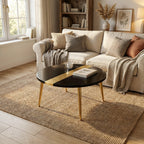 A living room scene featuring a black round coffee table with gold accents and legs. The table sits on a woven jute rug, with a cream-colored sofa, pillows, and a blanket in the background. A window and bookshelf are also visible.