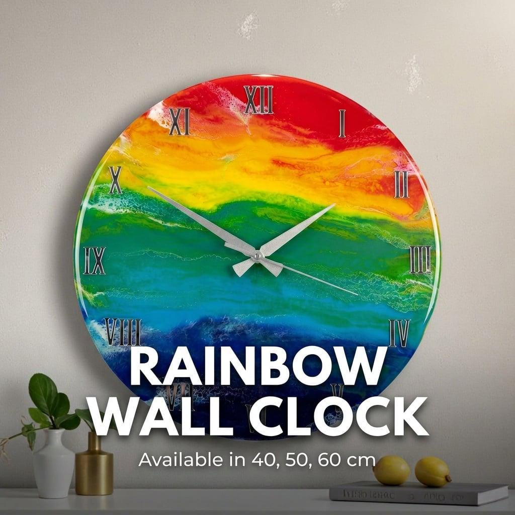 A round wall clock with a rainbow design. The clock face features a vibrant blend of red, orange, yellow, green, blue, and purple hues. Silver Roman numerals mark the hours, and silver hands indicate the time. The text "RAINBOW WALL CLOCK" is displayed on the clock face.