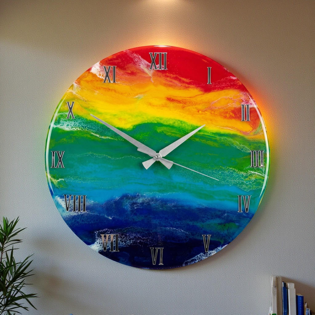 A round wall clock with a vibrant rainbow design. The clock face features a gradient of colors, including red, orange, yellow, green, blue, and indigo. Silver Roman numerals mark the hours, and silver hands indicate the time. The clock is mounted on a gray wall.