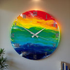 A round wall clock with a vibrant rainbow design. The clock face features a gradient of colors, including red, orange, yellow, green, blue, and indigo. Silver Roman numerals mark the hours, and silver hands indicate the time. The clock is mounted on a gray wall.