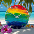 A round wall clock with a vibrant rainbow design. The clock face features a gradient of colors, from red at the top to blue at the bottom, with silver Roman numerals and hands. The clock is set against a beach backdrop with flowers.