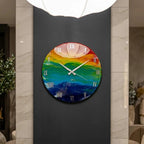 A round wall clock with a colorful, abstract design. The clock face features a rainbow of colors, with white Roman numerals and hands. The clock is mounted on a dark gray wall.