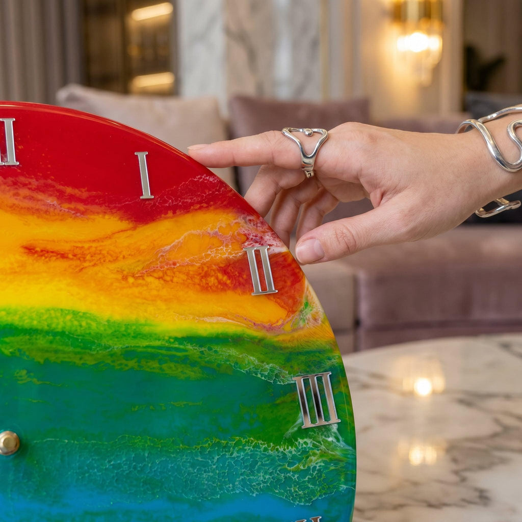 A colorful wall clock with a rainbow design. The clock face features Roman numerals and a gold-colored stud. The clock is being touched by a hand wearing a silver ring and bracelet.