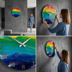 A round wall clock with a vibrant rainbow design. The clock face features a gradient of colors, including red, orange, yellow, green, and blue. Roman numerals mark the hours, and the hands are white.