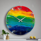 A round wall clock with a rainbow-colored face. The clock face features a blend of red, orange, yellow, green, blue, and indigo hues. Silver Roman numerals mark the hours, and silver hands indicate the time. The clock is mounted on a light gray wall.