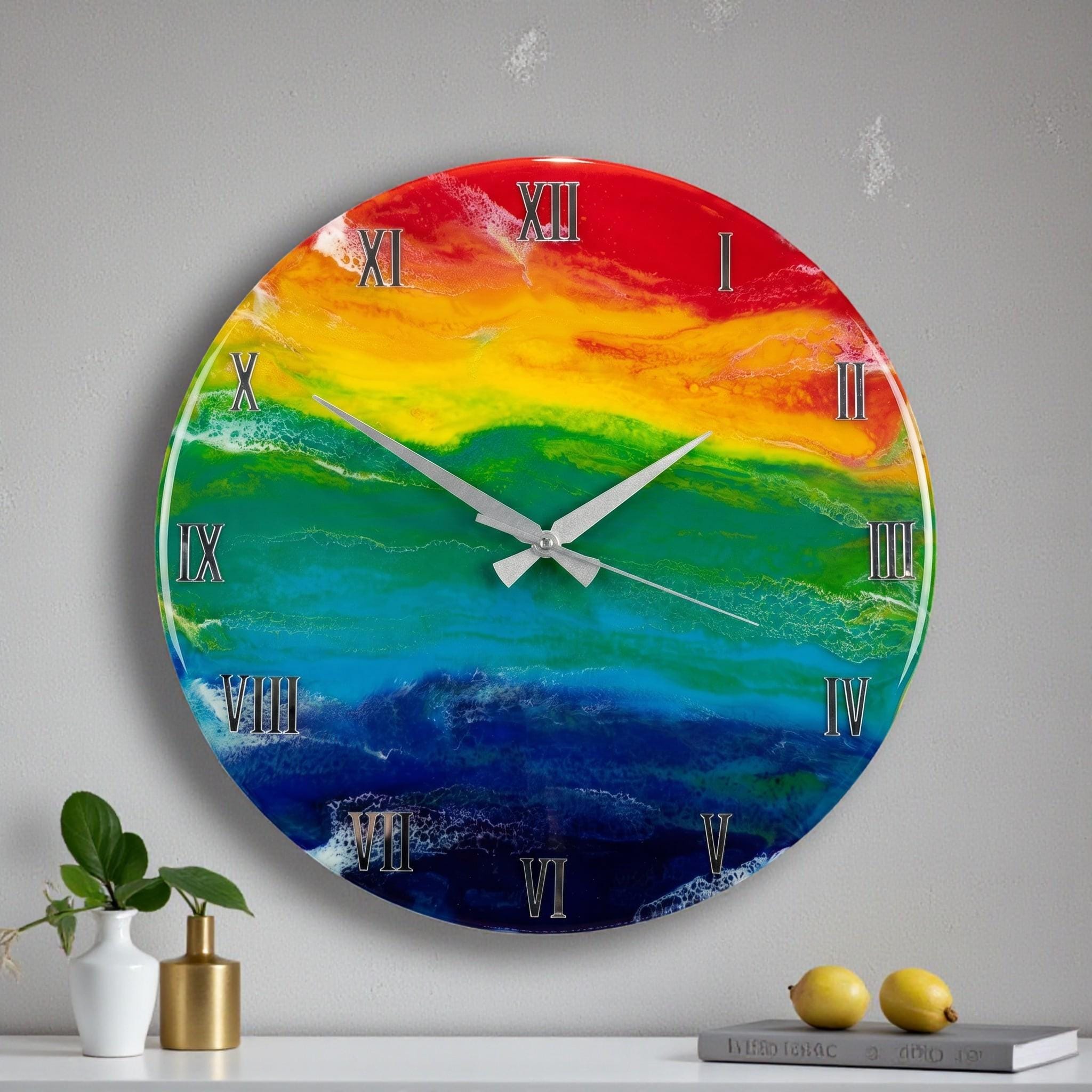 A round wall clock with a rainbow-colored face. The clock face features a blend of red, orange, yellow, green, blue, and indigo hues. Silver Roman numerals mark the hours, and silver hands indicate the time. The clock is mounted on a light gray wall.