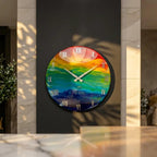 A round wall clock with a colorful, abstract design. The clock face features a rainbow of colors, with white Roman numerals and silver hands. The clock is mounted on a dark gray wall.