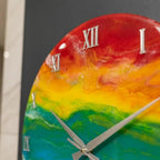 A round wall clock with a vibrant rainbow-colored face. The clock features silver Roman numerals and hands. The color transitions from red to orange, yellow, green, and blue. The clock is a decorative item.