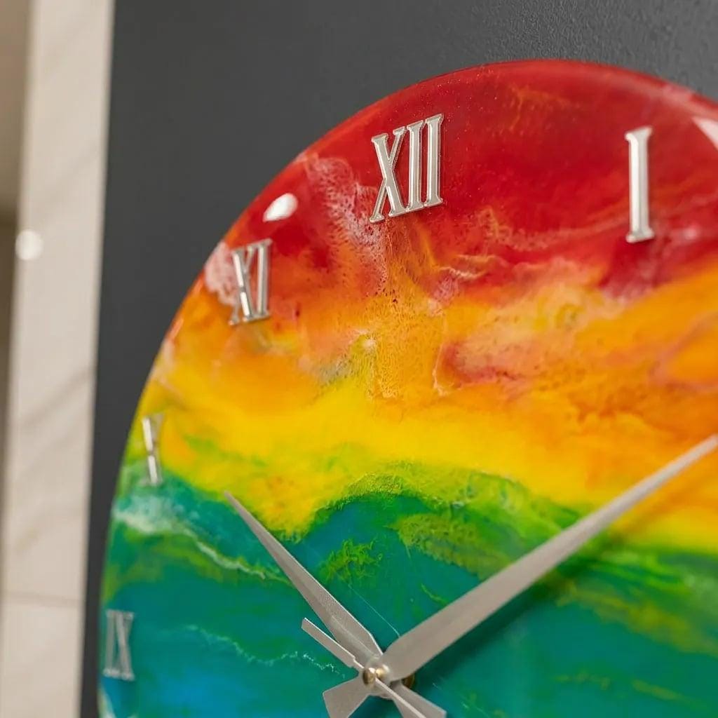 A round wall clock with a vibrant rainbow-colored face. The clock features silver Roman numerals and hands. The color transitions from red to orange, yellow, green, and blue. The clock is a decorative item.