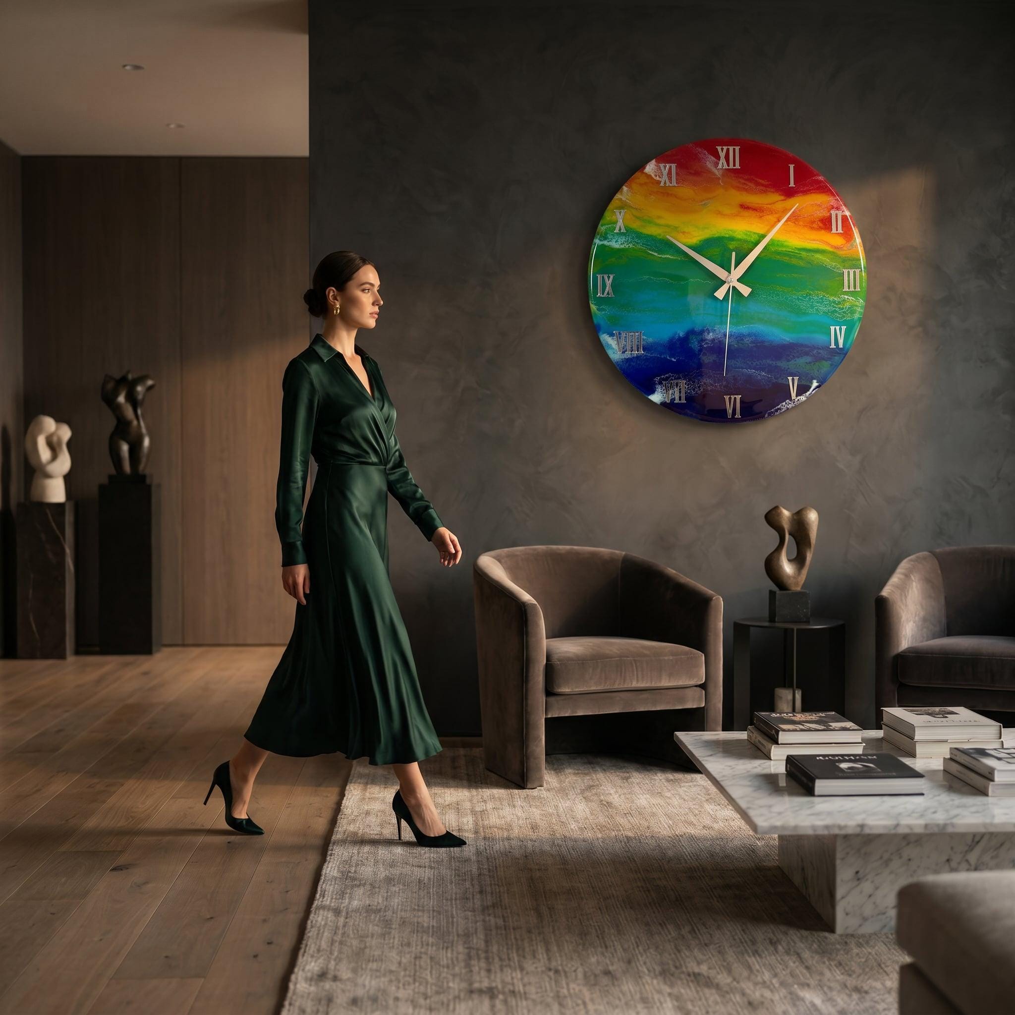 A large, round wall clock with a vibrant rainbow-colored design. The clock features silver-colored hands and Roman numeral hour markers. The clock is mounted on a dark gray wall in a modern living room setting.