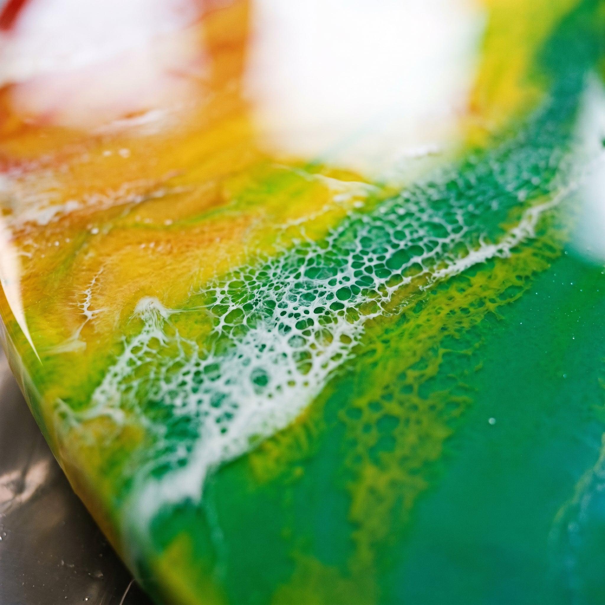 Close-up of a colorful resin art piece. The artwork features a blend of yellow, green, and white hues, with a textured, bubbly pattern along the edges. The colors transition smoothly, creating a vibrant and dynamic visual.