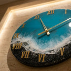 A round wall clock with a blue and white ocean-themed design. The clock face features gold Roman numerals and hands. The clock's design includes a black, textured border, and the clock is mounted on a beige wall.
