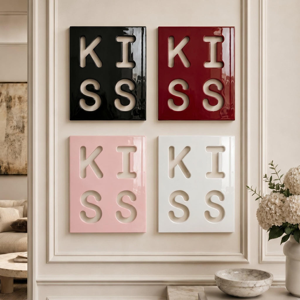 Four rectangular wall art panels arranged in a grid, each featuring the word KISS cut out in a bold, block font. The top left panel is glossy black, the top right is deep red, the bottom left is soft pink, and the bottom right is white.