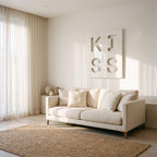 A cream-colored sofa with four matching throw pillows sits in a room with a large rectangular wall art piece featuring the word KISS in recessed lettering. A woven jute rug lies on the floor in front of the sofa.