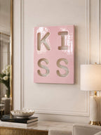 A rectangular, glossy, light pink wall art piece featuring the word KISS cut out in a bold, block-letter style. The letters are arranged in two rows of two, with K and I on the top row and S and S on the bottom row, creating a cutout effect.