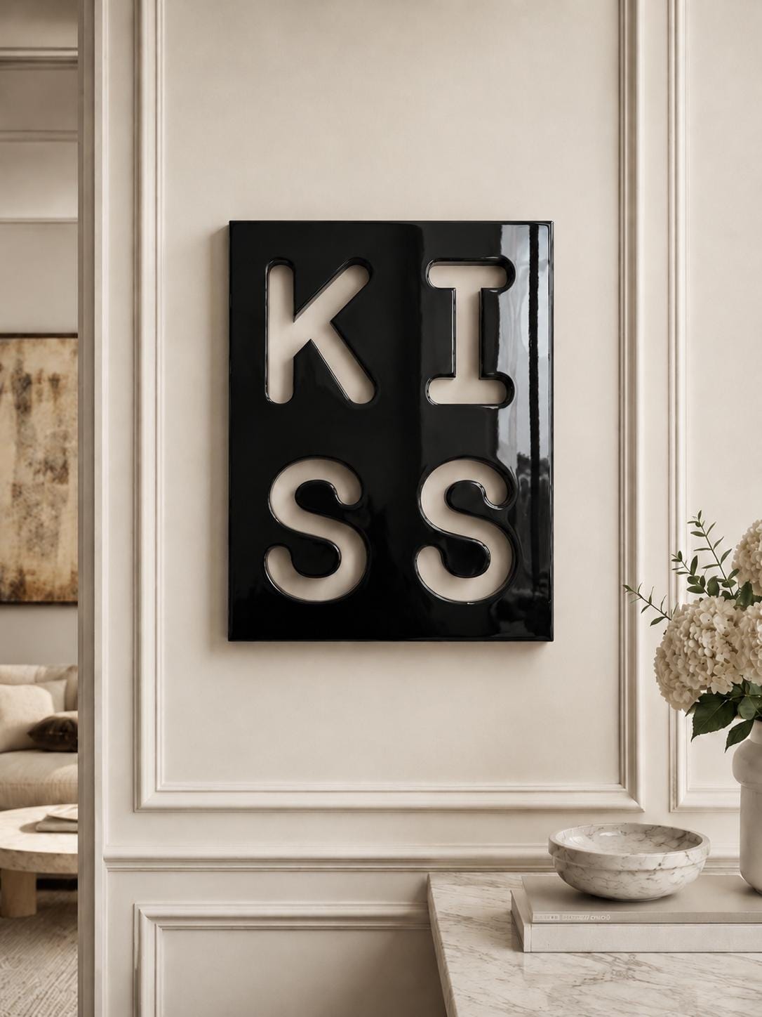 A rectangular black wall art piece featuring the word KISS in large, bold, white cut-out lettering. The surface of the black panel is glossy and reflective, creating a high-contrast visual effect against the white letters.