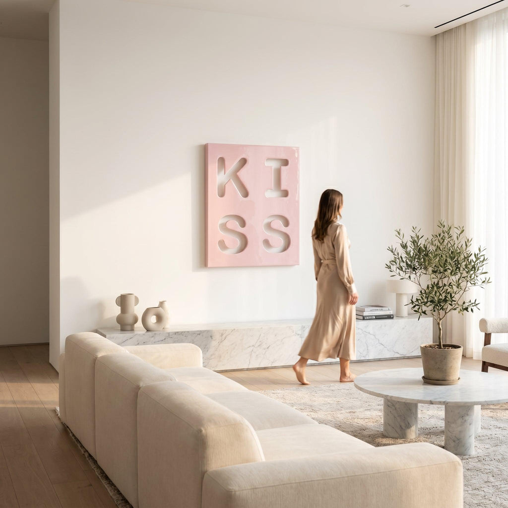 A square wall art piece featuring a light pink background with the word KISS cut out in a bold, blocky font. The letters are arranged in a two-by-two grid, with K and I on the top row and S and S on the bottom row, creating a cutout effect.