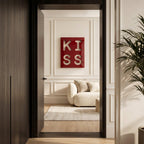 A square red wall art piece featuring the white cutout letters K, I, S, and S arranged in a two-by-two grid. The artwork is displayed on a white paneled wall above a cream-colored sofa in a room viewed through a dark doorway.