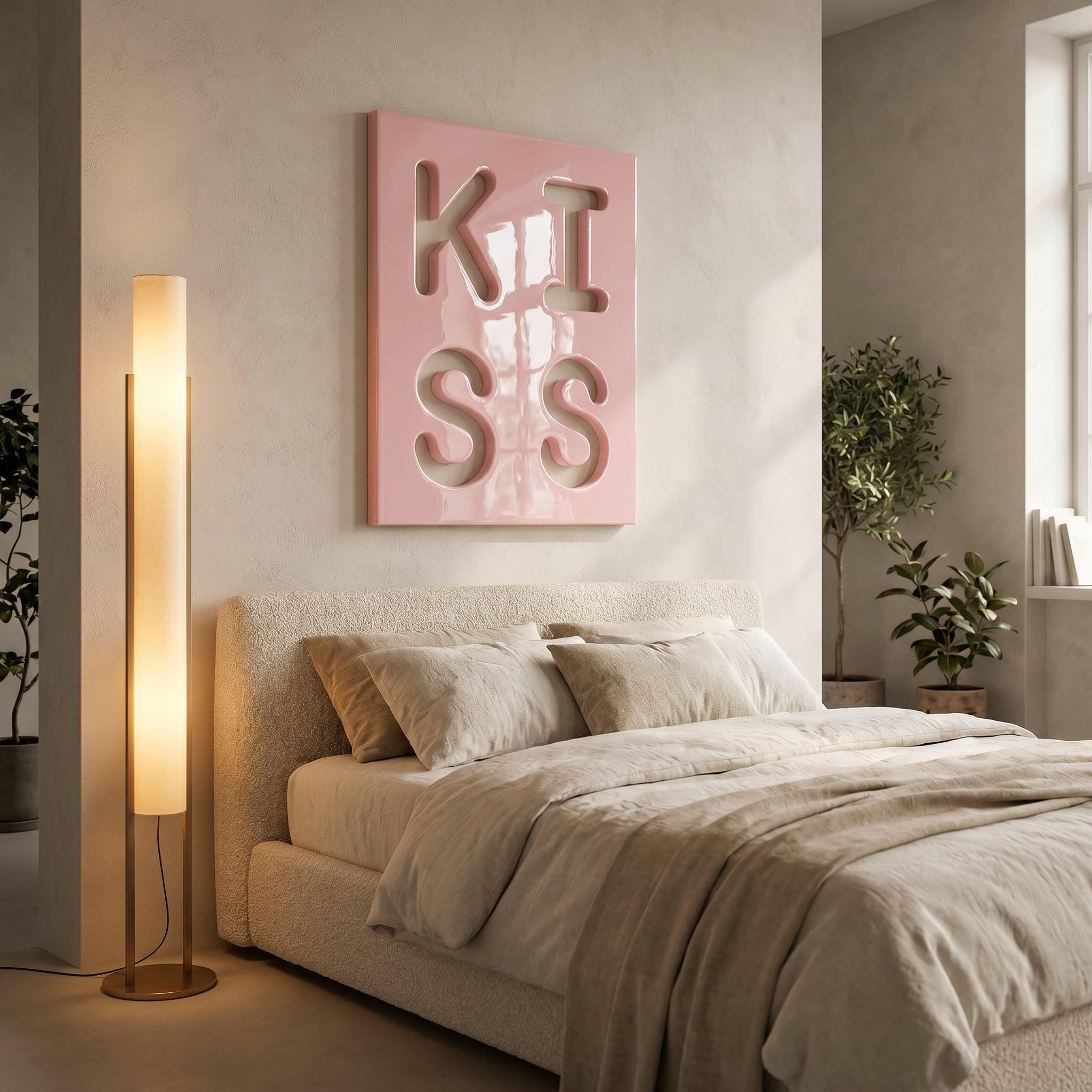 A rectangular wall art piece featuring the word KISS in large, cutout, three-dimensional letters against a glossy, light pink background. The artwork is mounted on a neutral-toned wall above a bed with beige bedding and pillows.