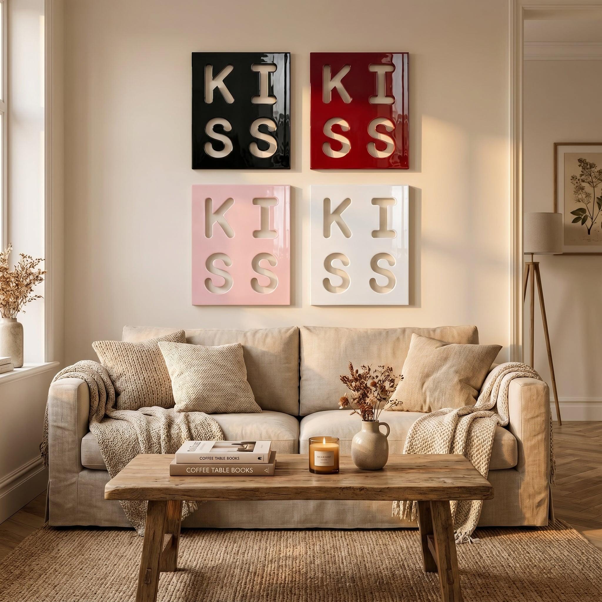 Four square wall art panels arranged in a grid, each featuring the word KISS in large, cut-out lettering. The top left panel is black, the top right is red, the bottom left is light pink, and the bottom right is white.