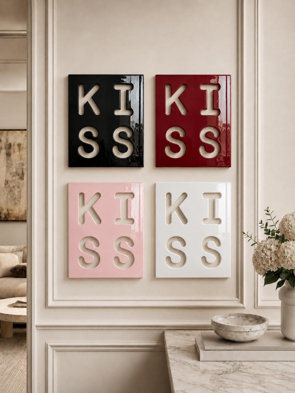 Four square wall art panels arranged in a grid, each featuring the word KISS cut out in a bold, block typeface. The panels are finished in glossy black, deep red, soft pink, and white, with the letters revealing the wall color behind.
