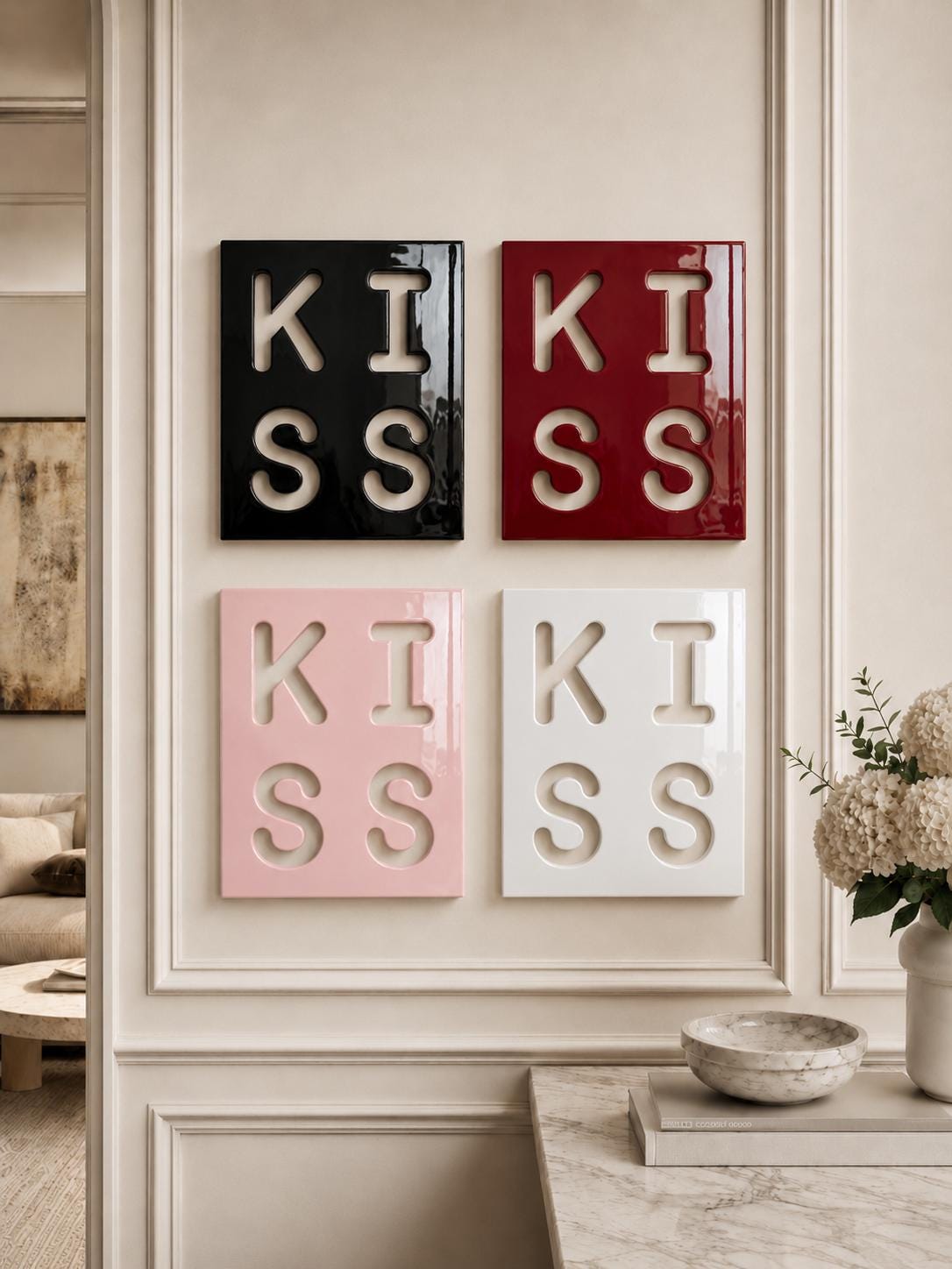 Four square wall art panels arranged in a grid, each featuring the word KISS cut out in a bold, block typeface. The panels are finished in glossy black, deep red, soft pink, and white, with the letters revealing the wall color behind.