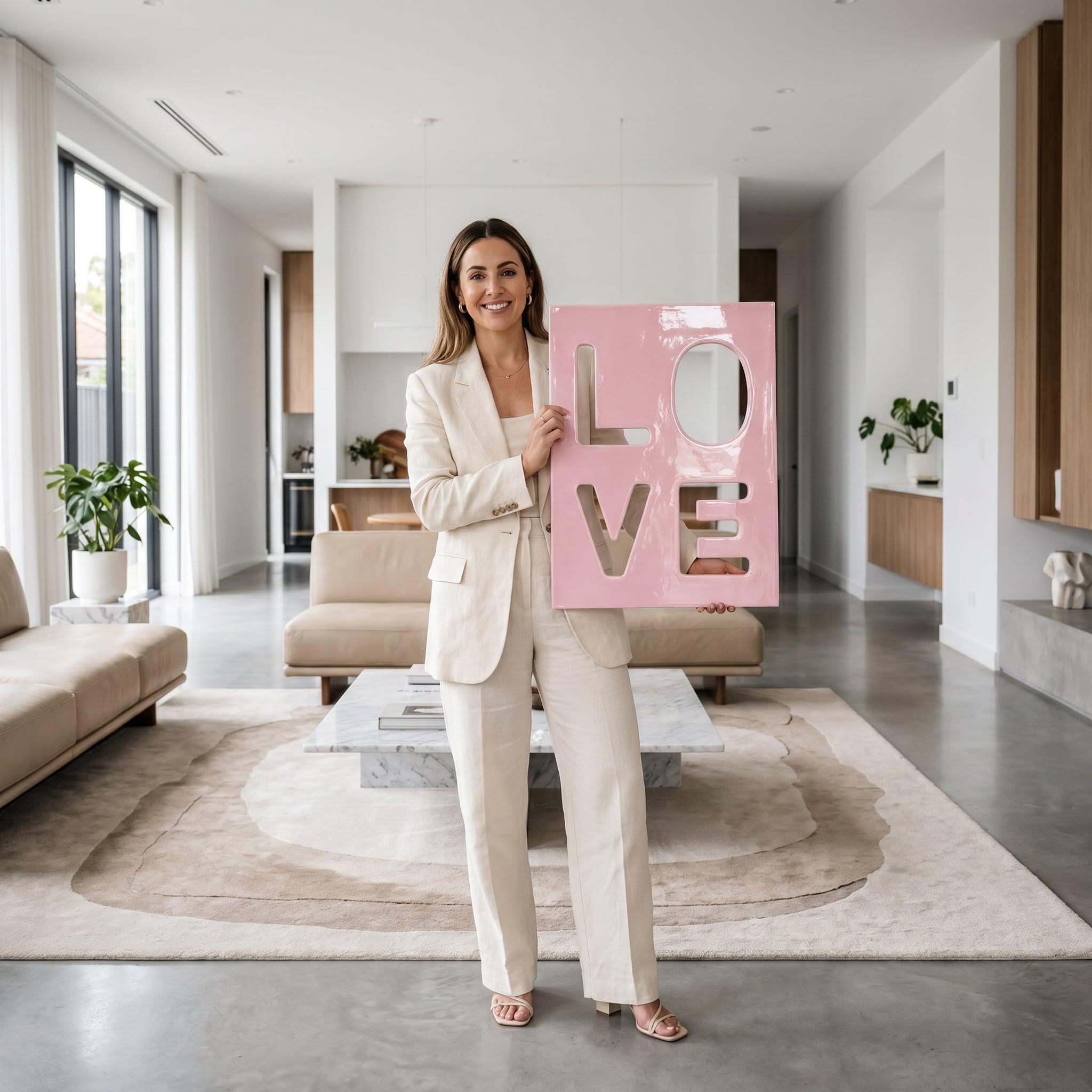 A rectangular light pink wall art piece featuring the word LOVE cut out in a bold, block-style font. The letters are negative space, allowing the background to be seen through the openings of the L, O, V, and E characters.