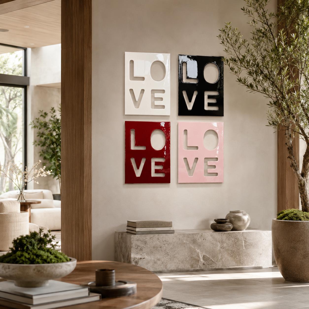 Four square wall art panels arranged in a grid, each featuring the word LOVE cut out in a bold, block-letter style. The panels are finished in glossy white, black, deep red, and soft pink, creating a modern, high-contrast display.