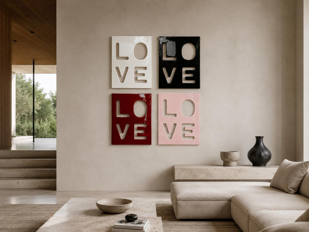 Four square wall art panels arranged in a grid, each featuring the word LOVE cut out in a bold, block-letter style. The panels are finished in glossy white, black, deep red, and soft pink, creating a high-contrast decorative display.