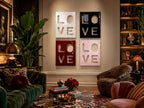Four square wall art panels arranged in a grid, each featuring the word LOVE in a cutout style. The top left panel is white, the top right is black, the bottom left is deep red, and the bottom right is a soft pastel pink.