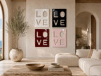 Four square wall art panels arranged in a grid, each featuring the word LOVE in a cutout style. The top left panel is white, the top right is black, the bottom left is deep red, and the bottom right is light pink, all with a glossy finish.