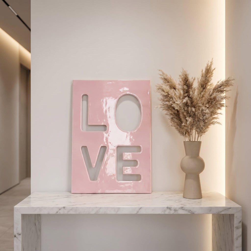 A rectangular pink plaque with the word LOVE cut out in a bold, block-letter style. Next to the plaque is a beige, rounded ceramic vase filled with tall, feathery, light brown dried pampas grass stems.