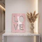 A rectangular pink plaque with the word LOVE cut out in a bold, block-letter style. Next to the plaque is a beige, rounded ceramic vase filled with tall, feathery, light brown dried pampas grass stems.