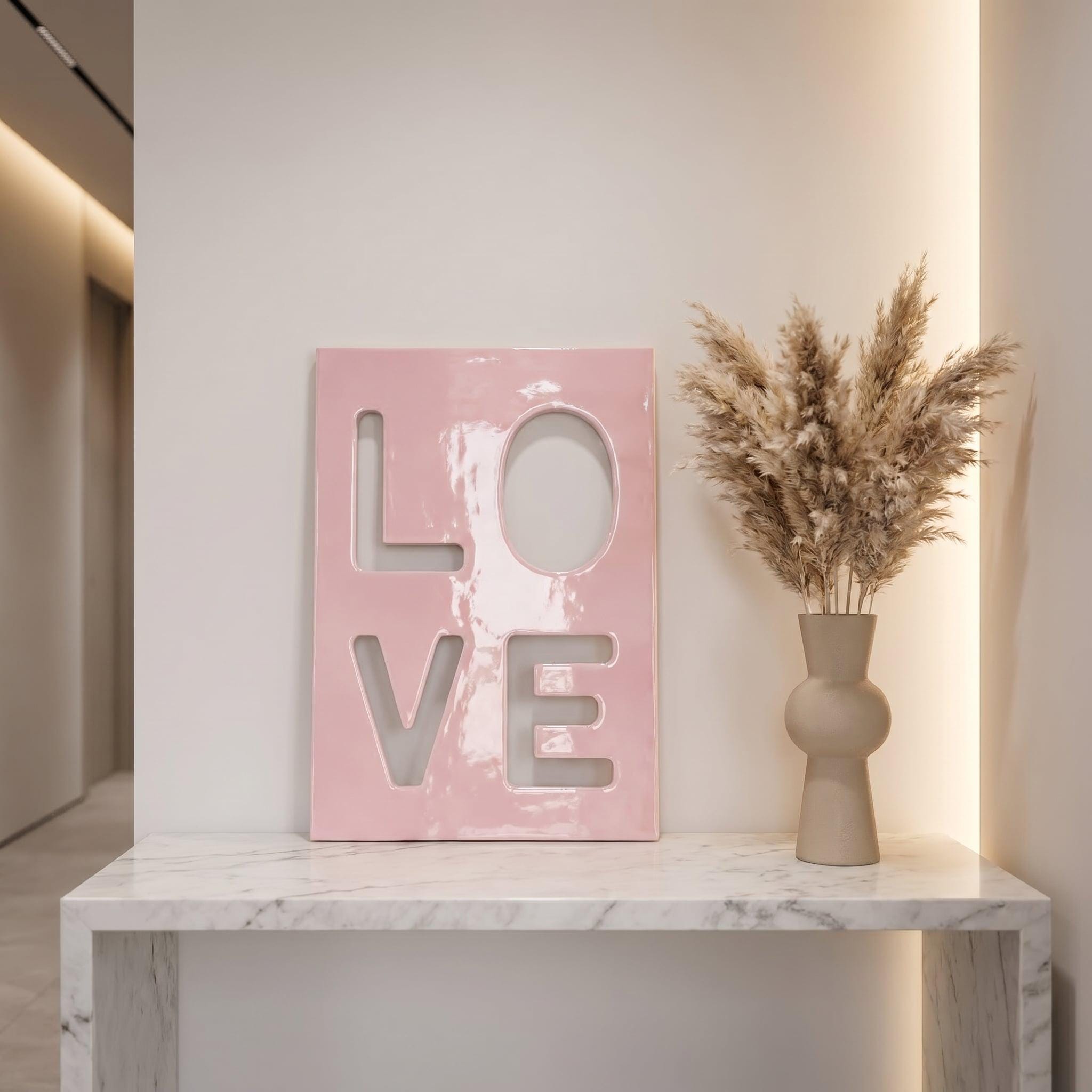 A rectangular pink plaque with the word LOVE cut out in a bold, block-letter style. Next to the plaque is a beige, rounded ceramic vase filled with tall, feathery, light brown dried pampas grass stems.