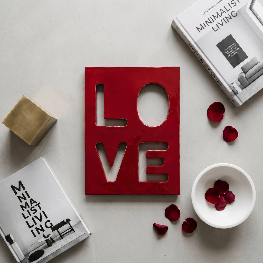 A rectangular red plaque with the word LOVE cut out in a block font. The plaque is surrounded by two books titled Minimalist Living, a small gold cube, a white bowl containing red rose petals, and loose red rose petals.