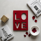 A rectangular red plaque with the word LOVE cut out in a block font. The plaque is surrounded by two books titled Minimalist Living, a small gold cube, a white bowl containing red rose petals, and loose red rose petals.