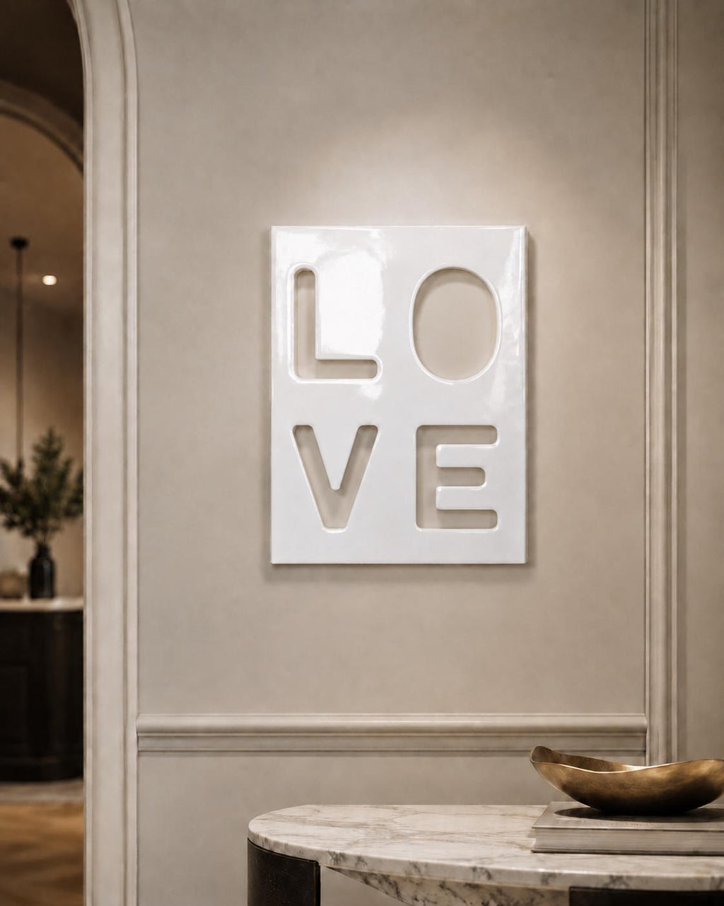 A rectangular white wall plaque featuring the word LOVE in a cut-out style. The letters are arranged in a two-by-two grid, with L and O on the top row and V and E on the bottom row. The surface of the plaque has a smooth, glossy finish.
