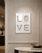 A rectangular white wall plaque featuring the word LOVE in a cut-out style. The letters are arranged in a two-by-two grid, with L and O on the top row and V and E on the bottom row. The surface of the plaque has a smooth, glossy finish.
