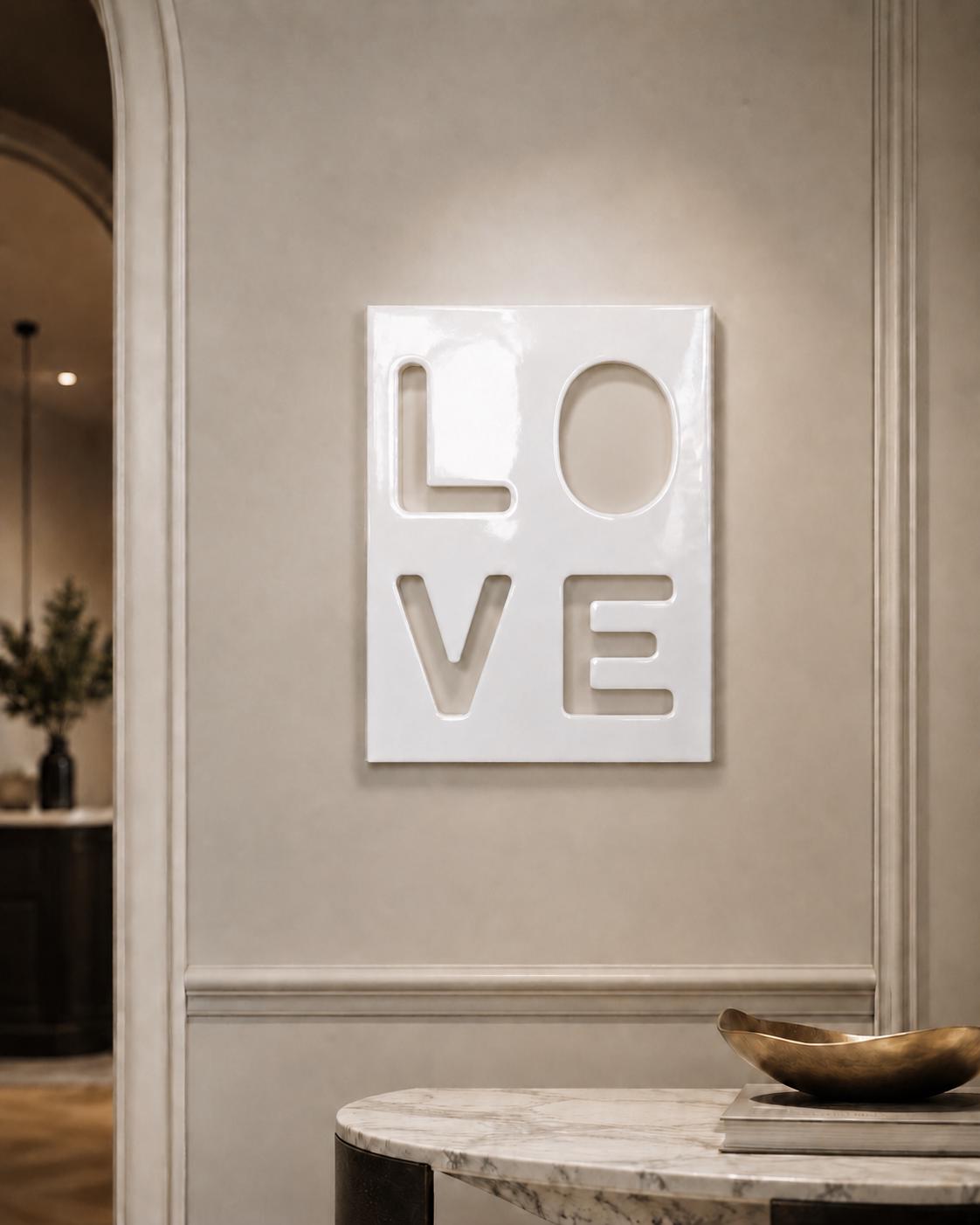 A rectangular white wall plaque featuring the word LOVE in a cut-out style. The letters are arranged in a two-by-two grid, with L and O on the top row and V and E on the bottom row. The surface of the plaque has a smooth, glossy finish.