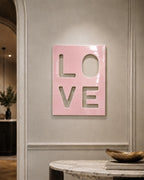 A rectangular wall art piece with a glossy light pink finish featuring the word LOVE cut out in a stacked two-by-two grid format. The letters are negative space, allowing the wall color to show through the cutouts in the panel.