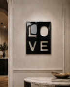 A rectangular black wall art piece featuring the word LOVE cut out in a bold, block-letter style. The glossy surface of the black panel contrasts with the negative space of the letters, which reveal the wall color behind the artwork.