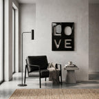 A black armchair with a textured throw blanket, a tall black floor lamp, and a small concrete side table holding books and a mug. On the wall hangs a black rectangular art piece featuring the white cutout letters L, O, V, and E.