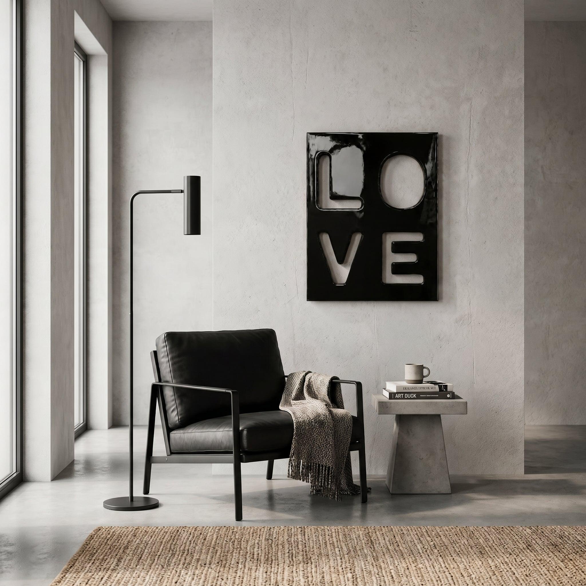 A black armchair with a textured throw blanket, a tall black floor lamp, and a small concrete side table holding books and a mug. On the wall hangs a black rectangular art piece featuring the white cutout letters L, O, V, and E.