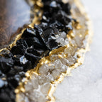 A decorative coaster featuring a dark brown center surrounded by a textured border of black and clear crystalline fragments. The edge is defined by a metallic gold finish that highlights the irregular, organic shape of the piece.