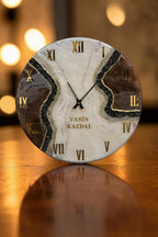 A round wall clock featuring a white and brown marbled face with gold Roman numerals. A jagged, textured black and gold glitter vein runs diagonally across the center. The text YASIN KAZDAL is printed in gold below the clock hands.